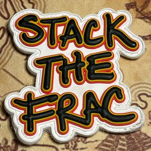 May include: A white, die-cut sticker with the words "STACK THE FRAC" in black, yellow, and red lettering. The text has a 3D effect with a black outline. The sticker is on a tan, textured surface.