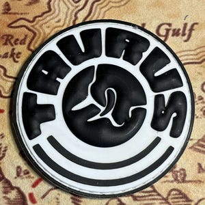 May include: A round, black and white emblem with the word "TAURUS" in bold, block letters. The center features a stylized black design. The emblem is set against a map background.