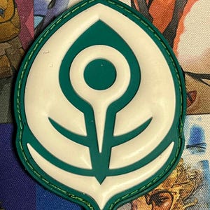 Può includere: Una toppa verde e bianca a forma di foglia. La toppa presenta un design bianco al centro, un bordo verde e un bordo cucito verde. Il design include un cerchio, una linea verticale e linee curve.