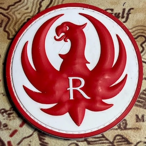 May include: A round, red and white patch featuring a stylized red phoenix with outstretched wings. The letter "R" is centered within the design. The patch has a white border and a red edge.