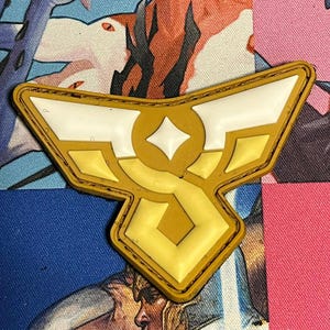 May include: A yellow and white emblem with a diamond shape in the center and wing-like shapes on the sides. The emblem has a gold border and is set against a colorful background with comic book art.