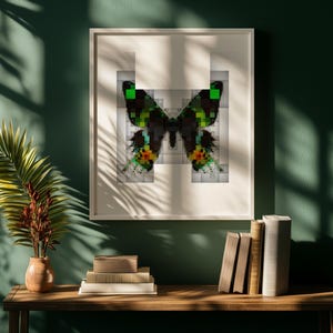 May include: A framed print of a pixelated butterfly in a white frame, set against a dark green wall. The butterfly design uses black, green, yellow, and orange. A wooden table displays books and a vase with decorative plants.