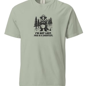 Op de afbeelding: Een saliegroen T-shirt met korte mouwen en een zwarte grafische print van een das met een hoed en rugzak in een bos. De tekst onder de das luidt, I'M NOT LOST. THIS IS A SHORTCUT. Het shirt heeft een ronde halslijn.