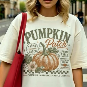 May include: An off-white short-sleeve t-shirt featuring a graphic of an orange pumpkin with the text PUMPKIN Patch, Fresh From The Patch, Farm Fresh, Locally Grown, and Pumpkins, Corn Maze, Apple Cider. A red tote bag hangs over the shoulder.