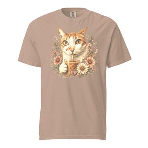 May include: A light tan short-sleeve t-shirt featuring a central graphic of a ginger tabby cat holding a clear plastic cup with an iced beverage and a straw, surrounded by a circular arrangement of various pink and white flowers and green foliage.