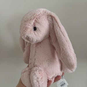 May include: A soft, light pink bunny toy with long, drooping ears. The plush rabbit has a fluffy texture and black eyes. A white tag is attached to the ear.