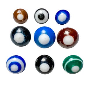 May include: A collection of nine spherical glass beads arranged in three rows. Each bead features a central white circular inlay surrounded by various colours including brown, black, blue, dark red, and green, with some displaying concentric ring patterns.