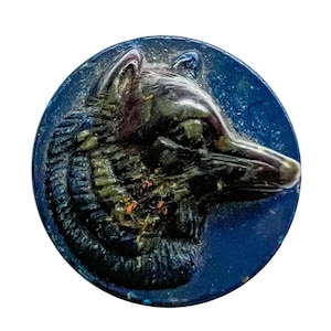 May include: A circular button featuring a raised, three-dimensional relief of a wolf head in profile. The button has a deep navy blue base color with metallic, dark green, and bronze-toned highlights accentuating the wolf's fur and features.
