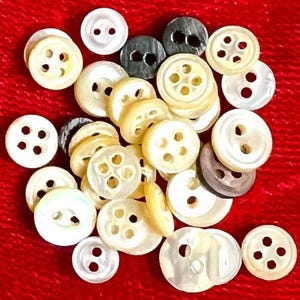 Può includere: Una collezione di bottoni assortiti in varie dimensioni e colori, tra cui crema, bianco e grigio. I bottoni hanno due o quattro fori e sono sparsi su una superficie rossa. I bottoni sono rotondi e realizzati in un materiale lucido.