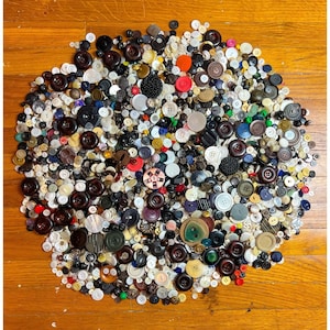May include: A large, dense collection of assorted sewing buttons in various sizes, shapes, and colors, including black, brown, white, red, green, and metallic tones, scattered in a circular arrangement on a wooden surface.