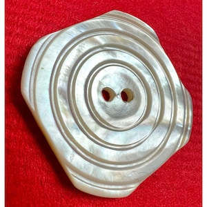 Könnte beinhalten: Ein großer, cremeweißer, irisierender Perlmuttknopf mit quadratischer Form und abgerundeten Ecken. Die Oberfläche weist konzentrische, kreisförmige Rillen auf, die von der Mitte ausgehen, welche zwei kleine, vertikal ausgerichtete Löcher enthält.