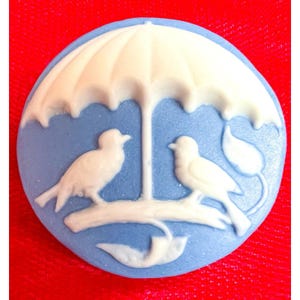 May include: A round, light blue cameo with a white umbrella and two birds perched on a branch. The cameo is set against a red background. The birds and umbrella are raised in relief.