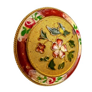 May include: A round, gold-toned decorative button with a floral design. The center features a textured gold surface with a flower and leaf motif in red, white, and green. The outer ring is red with a floral border.