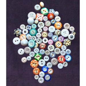 May include: A large collection of assorted sewing buttons in various shapes, sizes, and colors, including white, blue, orange, green, and patterned designs. The buttons feature different hole counts and textures, arranged in a cluster on a dark surface.