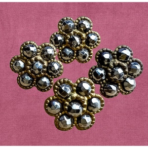 May include: Four antique-style decorative flower-shaped embellishments. Each flower has a gold-colored border and is filled with multiple silver-colored, faceted, round gems. The background is a solid, muted pink.