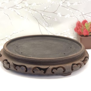 May include: A dark brown, round wooden display stand with a flat top and carved decorative accents. The stand has a low profile and is designed to hold a small object. The stand is made of wood and has a natural finish.