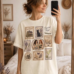 May include: An off-white t-shirt featuring a grid of postage stamp-style illustrations. The graphics include a house, a black cat, two people holding drinks labeled Midnight Margaritas, a book, lavender, a mortar and pestle, and candles.