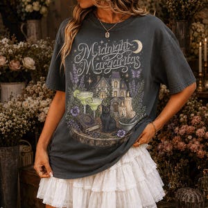 May include: A charcoal gray short-sleeve t-shirt featuring a graphic print with the text Midnight Margaritas, a house, a black cat, two margaritas, candles, and a book. The shirt is paired with a white tiered mini skirt with lace trim details.