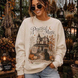 May include: A cream-colored crewneck sweatshirt featuring a graphic print of a Victorian house labeled The Owens House, a black cat, pumpkins, and autumn trees. The text reads Practical Magic and There's a little witch in all of us.