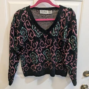 Può includere: Maglione nero con scollo a V con un motivo ripetuto di vortici rosa e verde acqua. Il maglione ha maniche lunghe e orlo e polsini a coste. Il marchio "Carducci" è visibile su un'etichetta vicino allo scollo. Il maglione è appeso a una gruccia rosa.