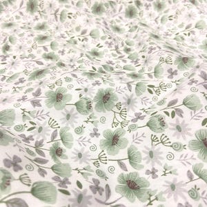 May include: Close-up of a white fabric with a repeating floral pattern. The design features sage green and grey flowers, leaves, and stems. The fabric has a soft, slightly wrinkled texture, suggesting it is a lightweight material.