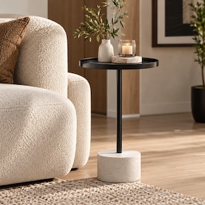 May include: A round black metal side table featuring a slender central post supported by a heavy cylindrical white marble base. The tabletop holds a small white ceramic vase with green foliage and a glass candle holder with a lit candle.