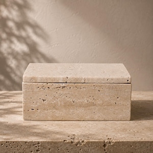 May include: A rectangular box crafted from beige travertine stone, featuring a natural porous texture and a flat, removable lid. The stone displays varied light tan and cream tones with visible surface indentations throughout the exterior.