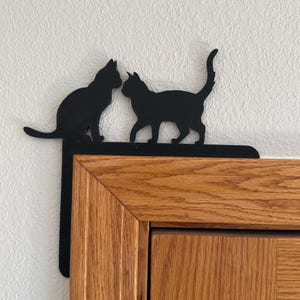 May include: Black metal cat silhouette decor featuring two cats. One cat is seated, the other is walking. The decor is mounted on a corner, with the cats facing each other. The background is a white wall and a wooden cabinet.