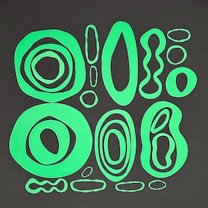 May include: A collection of bright neon green die-cut shapes arranged on a dark background. The set includes various circular, oval, and irregular organic forms, some featuring nested cutouts, creating a series of open frames and solid outlines.