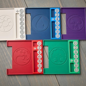 May include: A collection of six multi-colored game life counter trays. Each tray features a unique symbol and a numerical life counter from 0 to 12. The trays are white, blue, purple, red, and green.