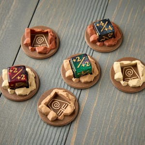 Puede incluir: Una colección de fichas y dados de juego. Las fichas son marrones con un diseño tallado y un borde rocoso. Los dados son multicolores con números blancos. Los artículos están dispuestos sobre una superficie de madera gris.