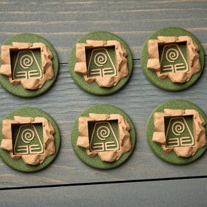 May include: Six green, circular tokens with a beige, stone-like border. Each token features a beige symbol within a square frame. The tokens are arranged on a gray-blue wooden surface.