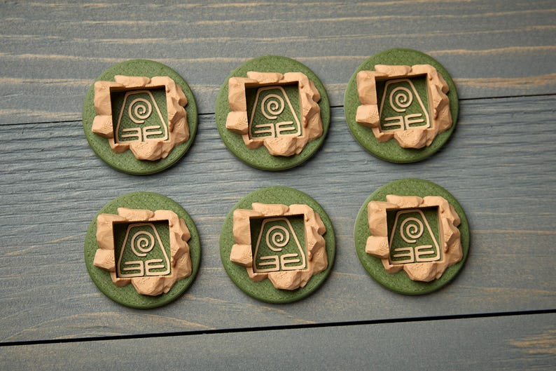 May include: Six green, circular tokens with a beige, stone-like border. Each token features a beige symbol within a square frame. The tokens are arranged on a gray-blue wooden surface.