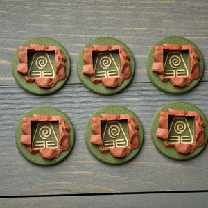 May include: Six round, green and brown decorative items with a gold-colored symbol. The items have a circular green base with a brown, rocky border. The gold symbol is a square with a spiral and horizontal lines.