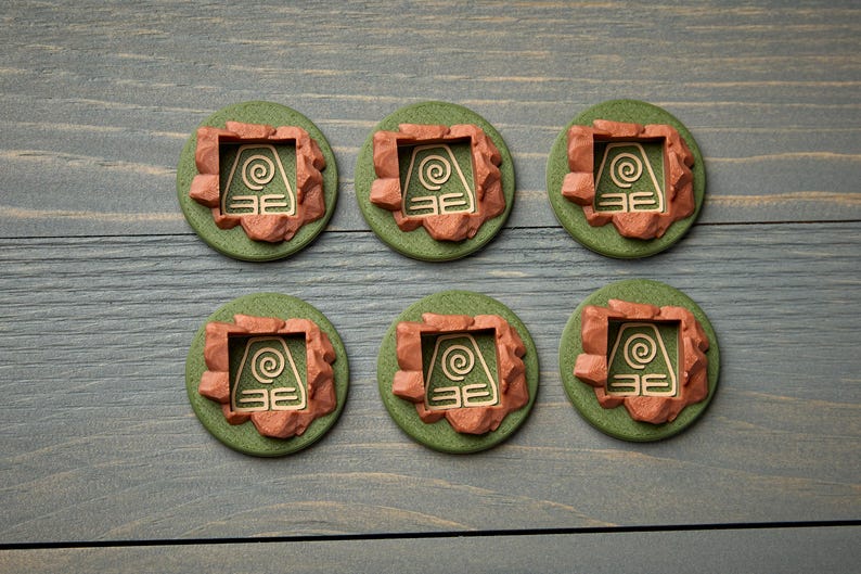 May include: Six round, green and brown decorative items with a gold-colored symbol. The items have a circular green base with a brown, rocky border. The gold symbol is a square with a spiral and horizontal lines.