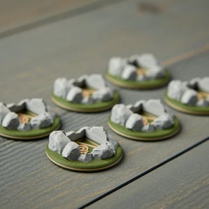 May include: Six miniature, circular terrain pieces. Each piece features a gray stone structure with a central opening, surrounded by green and tan details. The pieces are arranged on a gray wooden surface.
