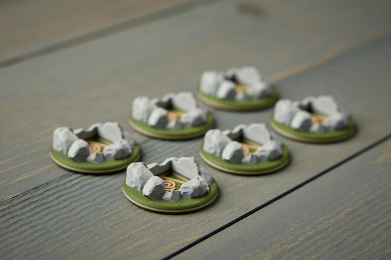 May include: Six miniature, circular terrain pieces. Each piece features a gray stone structure with a central opening, surrounded by green and tan details. The pieces are arranged on a gray wooden surface.