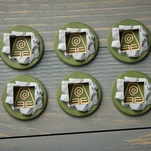 May include: Six round, green and gray decorative pieces with a gold-colored symbol. Each piece features a gold symbol within a gray stone-like frame, set on a green base. The pieces are arranged on a gray wooden surface.