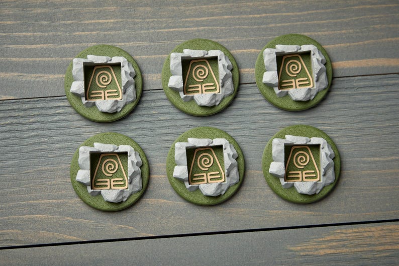 May include: Six round, green and gray decorative pieces with a gold-colored symbol. Each piece features a gold symbol within a gray stone-like frame, set on a green base. The pieces are arranged on a gray wooden surface.