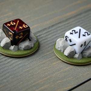 May include: Two dice, one black and one white, each resting on a small, round base with a green border and gray rocks. The black die has gold numbers, while the white die has black numbers. The dice are for tabletop games.