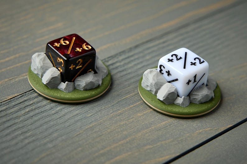 May include: Two dice, one black and one white, each resting on a small, round base with a green border and gray rocks. The black die has gold numbers, while the white die has black numbers. The dice are for tabletop games.