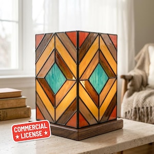 May include: A geometric stained glass lamp with a wooden base. The lamp features a pattern of orange, yellow, brown, and teal glass panels. The lamp is rectangular and sits on a dark wooden base. The lamp is on a light-coloured table.
