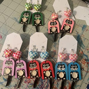 May include: A collection of handmade earrings featuring a cat design. Each pair has a different color scheme, including green, pink, purple, red, and blue. The earrings have a heart-shaped top and a black cat with a clock face.