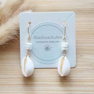 May include: Handmade earrings with white seashell pendants and small white beads, hanging from gold-coloured hoops. The earrings are presented on a white card with the text "Shellbeach.Ave HANDMADE JEWELLERY".