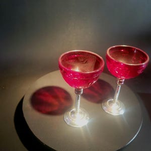 May include: Two ruby red champagne coupe glasses with textured bowls and silver stems. The glasses are displayed on a dark gray circular surface, casting a red shadow. The stem has a spiral design.