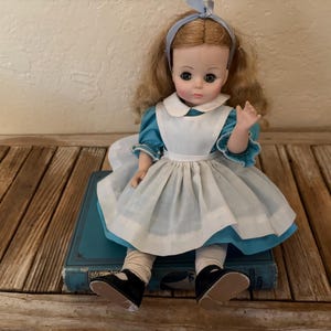 May include: A vintage doll dressed in a blue and white dress, seated on a blue book. The doll has blonde hair, a blue headband, and black shoes. The doll's dress has a white apron and a ruffled collar.