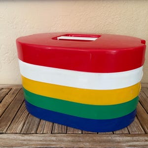 May include: A colorful, oval-shaped storage container with a red top, white, yellow, green, and blue bands. The container sits on a wooden surface. The top has a rectangular opening.