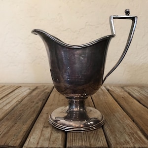 May include: Silver creamer with a rounded body and a single handle. The creamer has a pedestal base.