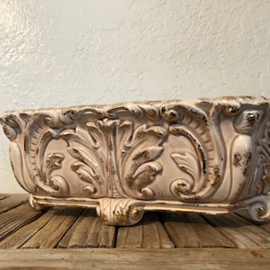 May include: A white ceramic planter with gold accents and an ornate design. The planter has a rectangular shape with four feet and a slightly curved top.