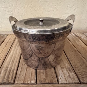 May include: A silver-colored ice bucket with a lid and two handles. The bucket has a decorative pattern on the lower half and sits on a wooden surface. The lid has a small knob.